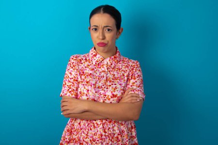 Photo for Gloomy dissatisfied woman wearing floral dress over blue studio background looks with miserable expression at camera from under forehead, makes unhappy grimace - Royalty Free Image