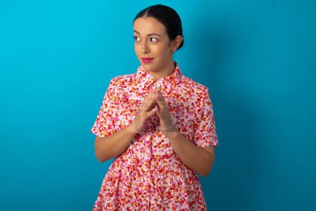 Téléchargez les photos : Femme portant une robe florale sur fond bleu studio doigts raides et semble mystérieux de côté a grand plan mal à l'esprit - en image libre de droit