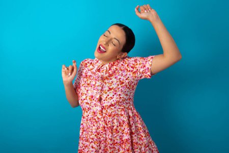 Téléchargez les photos : Photo de femme optimiste portant une robe florale sur fond bleu studio a du plaisir et danse vêtements insouciants être d'humeur parfaite fait des mouvements. Passe du temps libre sur disco partie - en image libre de droit