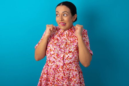 Photo for Woman wearing floral dress over blue studio background clenches fists and awaits for something nice happened looks away bites lips and waits announcement of results - Royalty Free Image