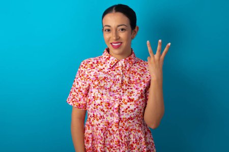 Photo for Woman wearing floral dress over blue studio background smiling and looking friendly, showing number three or third with hand forward, counting down - Royalty Free Image