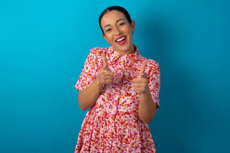 Foto de Mujer con vestido floral sobre fondo azul estudio dirige los dedos a la cámara selecciona a alguien. Te recomiendo. La mejor opción - Imagen libre de derechos