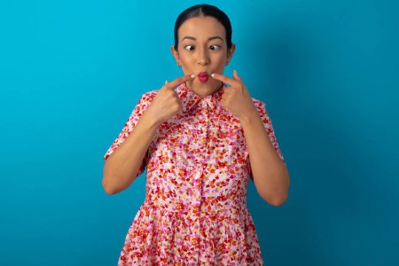 Téléchargez les photos : Femme portant une robe florale sur fond bleu studio croise les yeux et rend les lèvres de poisson grimace drôle - en image libre de droit