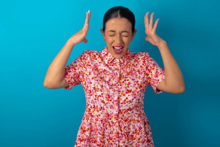 Photo for Woman wearing floral dress over blue studio background goes crazy as head goes around feels stressed because of horrible situation - Royalty Free Image