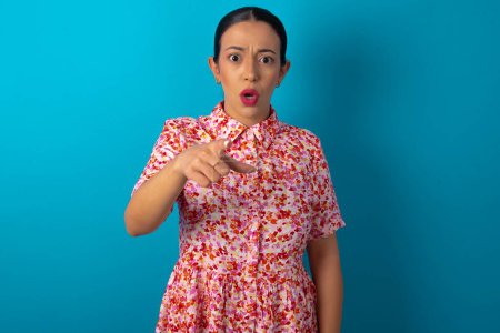 Téléchargez les photos : Femme choquée portant une robe florale sur fond bleu studio points à vous avec une expression stupéfaite - en image libre de droit