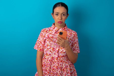 Foto de Mujer molesta insatisfecha con vestido floral utiliza aplicación de software móvil y navega información en Internet, sostiene la mano móvil moderna - Imagen libre de derechos