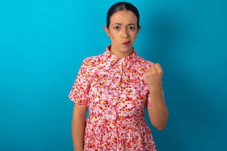 Photo for Woman wearing floral dress shows fist has annoyed face expression going to revenge or threaten someone makes serious look. I will show you who is boss - Royalty Free Image