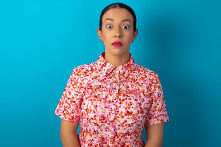 Photo for Stunned woman wearing floral dress over blue studio background stares reacts on shocking news. Astonished MODEL holds breath - Royalty Free Image