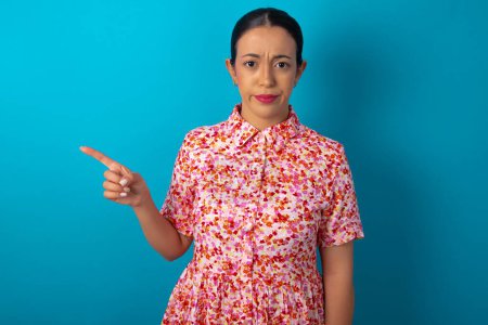 Photo for Serious woman wearing floral dress over blue studio background smirks face points away on copy space shows something unpleasant. Look at this advertisement. Big price concept - Royalty Free Image