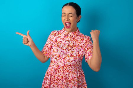 Téléchargez les photos : Femme joyeuse portant une robe florale sur fond de studio bleu montrant l'espace de copie annonce célébrant la chance - en image libre de droit