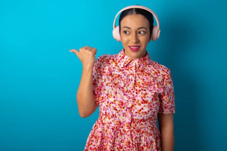 Téléchargez les photos : Femme portant une robe florale sur fond bleu studio écoute la piste audio via casque sans fil points pouce loin annonce l'espace de copie - en image libre de droit