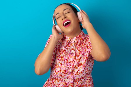 Photo for Woman wearing floral dress over blue studio background smiles broadly feels very glad listens favourite music track via wireless headphones closes eyes. - Royalty Free Image