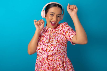 Photo for Carefree woman wearing floral dress over blue studio background with toothy smile raises arms dances carefree moves with rhythm of music listens music from playlist via headphones - Royalty Free Image