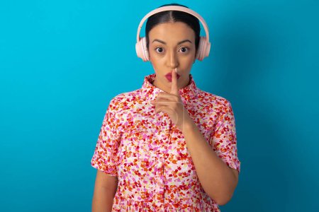 Photo for Woman wearing floral dress over blue studio background making hush gesture with finger on her lips wearing  wireless headphones. Be quiet. - Royalty Free Image