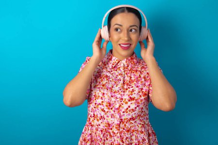 Photo for Joyful woman wearing floral dress over blue studio background sings song keeps hand near mouth as if microphone listens favorite playlist via headphones - Royalty Free Image
