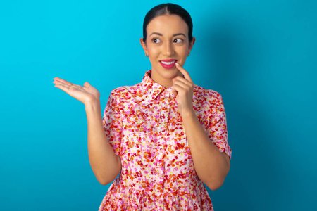 Foto de Mujer positiva con vestido floral sobre fondo azul estudio anuncio promocional toca los dientes con el dedo. - Imagen libre de derechos