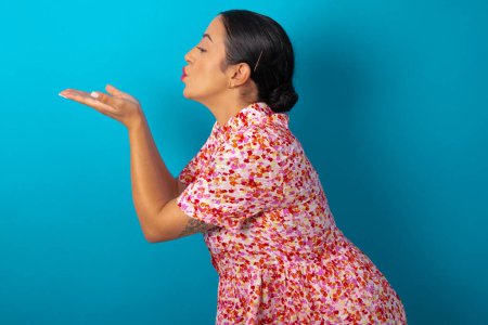 Téléchargez les photos : Profil vue de côté portrait de femme attrayante portant une robe florale sur fond bleu studio envoyer baiser d'air - en image libre de droit