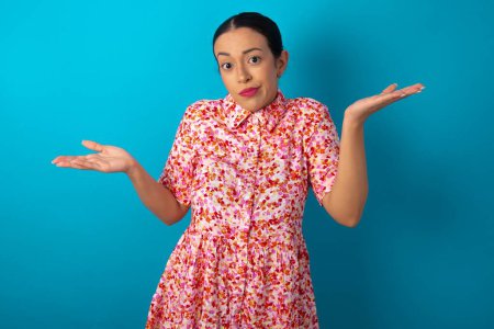 Photo for Careless attractive woman wearing floral dress over blue studio background shrugging shoulders, oops. - Royalty Free Image