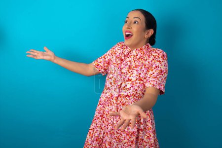 Téléchargez les photos : Drôle de femme étonnée portant une robe florale sur fond bleu studio regarder espace vide avec les bras ouverts prêt à attraper quelque chose. - en image libre de droit