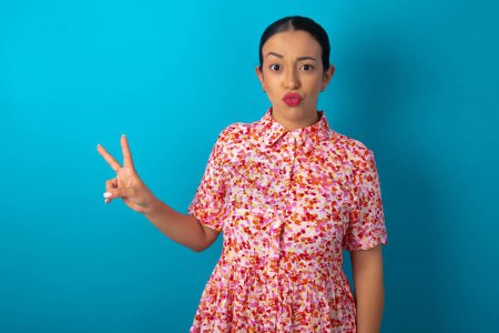 Téléchargez les photos : Femme portant une robe florale sur fond bleu studio fait geste de paix garde les lèvres pliées montre v signe. Concept de langage corporel - en image libre de droit