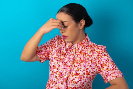 Photo for Sad woman wearing floral dress over blue studio background suffering from headache holding hand on her face - Royalty Free Image