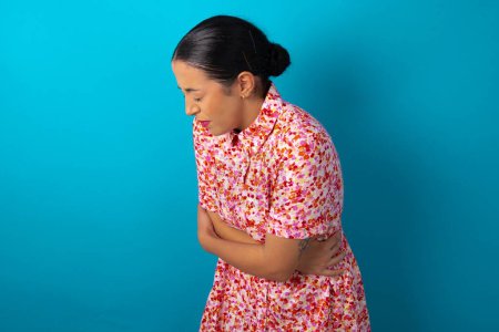 Téléchargez les photos : Femme portant une robe florale sur fond bleu studio a mal à l'estomac sur fond bleu - en image libre de droit