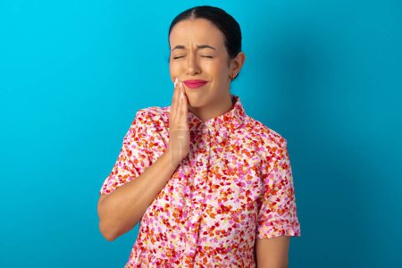 Photo for Woman wearing floral dress over blue studio background with toothache on blue background - Royalty Free Image
