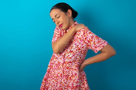 Téléchargez les photos : Femme portant une robe florale sur fond bleu studio a mal au dos - en image libre de droit