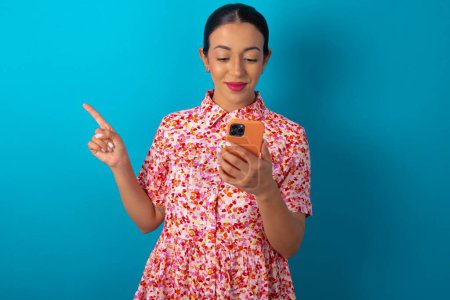 Téléchargez les photos : Femme souriante portant une robe florale sur fond bleu studio pointant du doigt l'espace vide tenant le téléphone dans une main - en image libre de droit