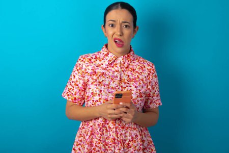Foto de Foto de mujer loca asombrada con vestido floral sobre fondo de estudio azul mantenga el concepto de retroalimentación de desagrado de teléfonos inteligentes - Imagen libre de derechos