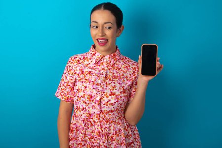 Photo for Photo of nice pretty woman wearing floral dress over blue studio background demonstrate phone screen hold hair tails - Royalty Free Image