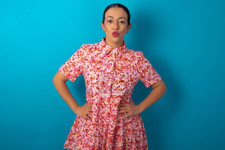 Téléchargez les photos : Portrait de belle femme drôle portant une robe florale sur fond bleu studio envoyant baiser d'air - en image libre de droit