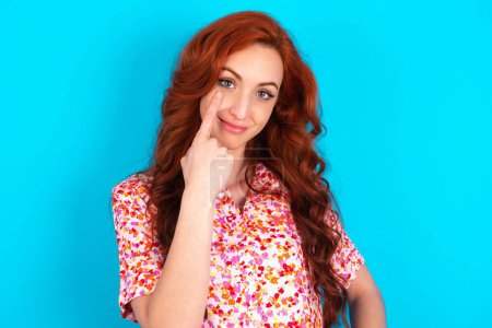 Photo for Redhead woman wearing floral dress over blue background looking, observing, keeping an eye on an object in front, or watching out for something. - Royalty Free Image