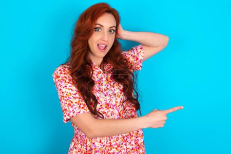 Photo for Redhead woman wearing floral dress over blue background feeling positive has amazed expression, indicates something. One hand on head and pointing with other hand. - Royalty Free Image