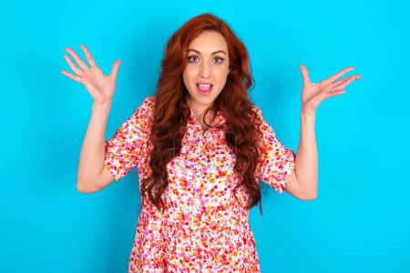 Photo for Redhead woman wearing floral dress over blue background raising hands up, having eyes full of happiness rejoicing his great achievements. Achievement, success concept. - Royalty Free Image