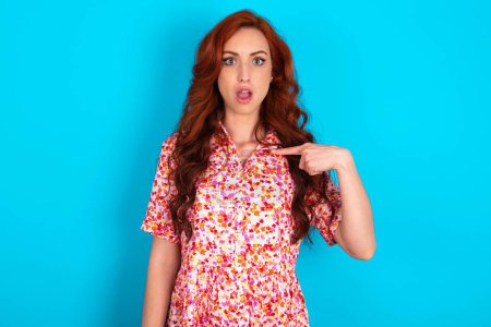 Photo for Redhead woman wearing floral dress over blue background being in stupor shocked, has astonished expression pointing at oneself with finger saying: Who me? - Royalty Free Image