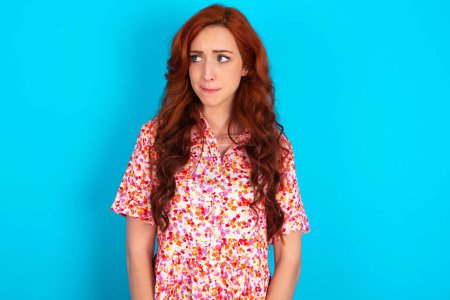 Photo for Amazed puzzled redhead woman wearing floral dress over blue background curves lips and has worried look, sees something awful in front. - Royalty Free Image