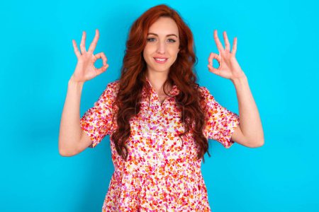Photo for Glad redhead woman wearing floral dress over blue background shows ok sign with both hands as expresses approval, has cheerful expression, being optimistic. - Royalty Free Image