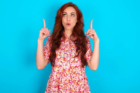 Photo for Redhead woman wearing floral dress over blue background being amazed and surprised looking and pointing up with fingers showing something strange. - Royalty Free Image