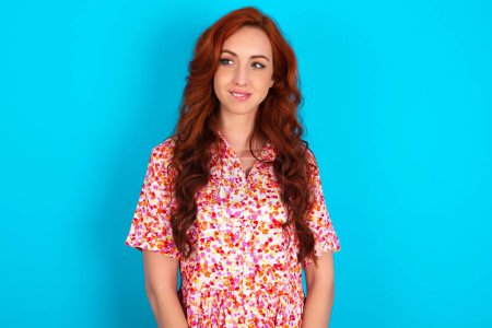 Photo for Redhead woman wearing floral dress over blue background with thoughtful expression, looks away keeps hands down bitting his lip thinks about something pleasant. - Royalty Free Image