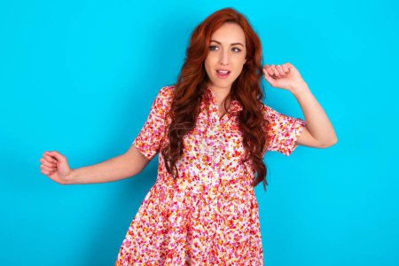 Photo for Redhead woman wearing floral dress over blue background dancing and listening music with headphones. - Royalty Free Image