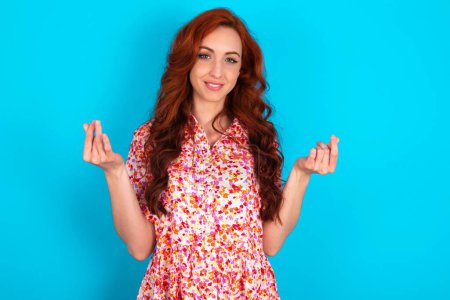 Photo for Redhead woman wearing floral dress over blue background making money gesture. - Royalty Free Image