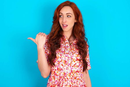 Photo for Stupefied redhead woman wearing floral dress over blue background with surprised expression, opens eyes and mouth widely, points aside with thumb, shows something strange. Advertisement concept. - Royalty Free Image