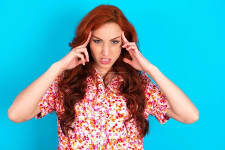 Photo for Redhead woman wearing floral dress over blue background concentrating hard on an idea with a serious look, thinking with both index fingers pointing to forehead. - Royalty Free Image