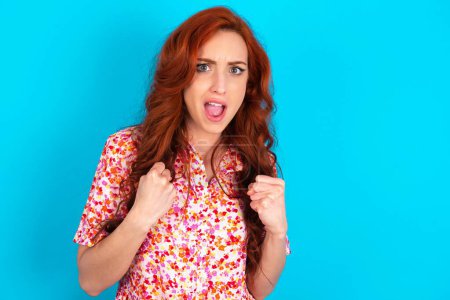 Photo for Redhead woman wearing floral dress over blue background rejoicing success and victory clenching his fists with joy being happy to achieve her aim and goals. Positive emotions, feelings. - Royalty Free Image