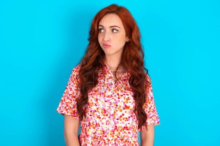 Photo for Redhead woman wearing floral dress over blue background looks pensively aside, plans actions after university, imagines what to do Thinks over about new project. - Royalty Free Image