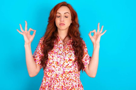 Photo for Redhead woman wearing floral dress over blue background doing yoga, keeping eyes closed, holding fingers in mudra gesture. Meditation, religion and spiritual practices. - Royalty Free Image