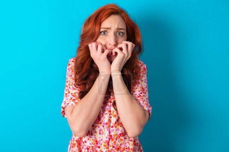 Anxiety - redhead woman wearing floral dress over blue background covering his mouth with hands scared from something or someone bitting nails.