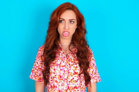 Photo for Body language. Disgusted stressed out redhead woman wearing floral dress over blue background frowning face, demonstrating aversion to something. - Royalty Free Image