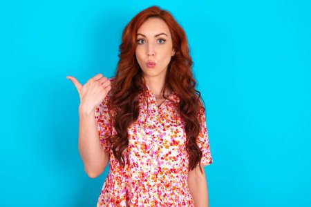 Photo for Shocked redhead woman wearing floral dress over blue background points with thumb away, indicates something. Check this out. Advertisement concept. - Royalty Free Image
